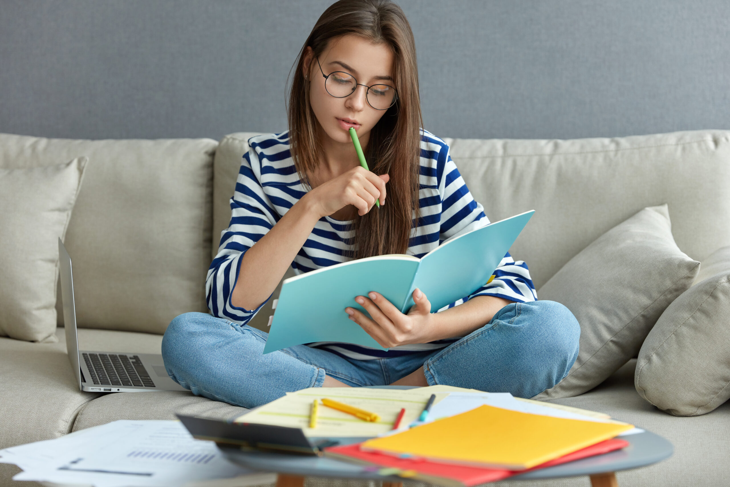 Studying online concept. Serious young woman being busy with remote freelance project, sits at comfortable sofa, writes notes, holds textbook, use laptop computer at home with wireless internet